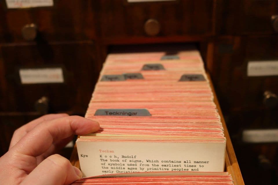 A hand leafing through typewritten index cards in an open library catalogue drawer, with a wall of wooden drawers behind it