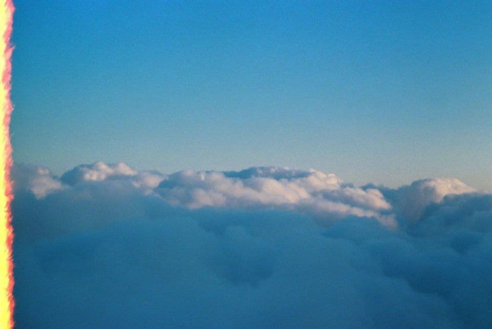 Film photograph taken above an unbroken layer of white clouds under a clear blue sky, with a red and yellow light leak running down the left edge
