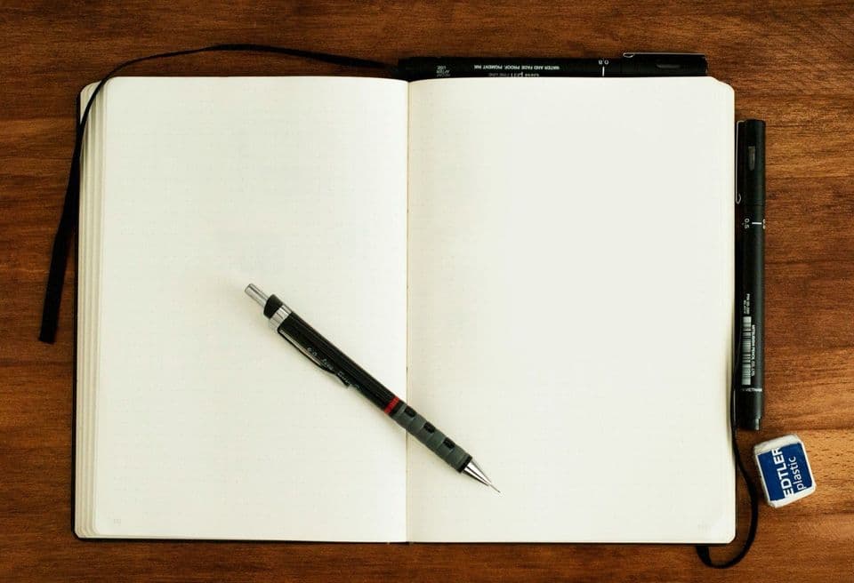 An open notebook with blank dot-grid pages on a dark wooden desk, a mechanical pencil lying across the spread and two fineliners beside it