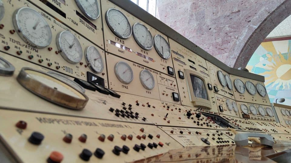 A vintage control room console photographed from below, dozens of round analogue dials with Cyrillic labels above rows of switches and coloured indicator lamps