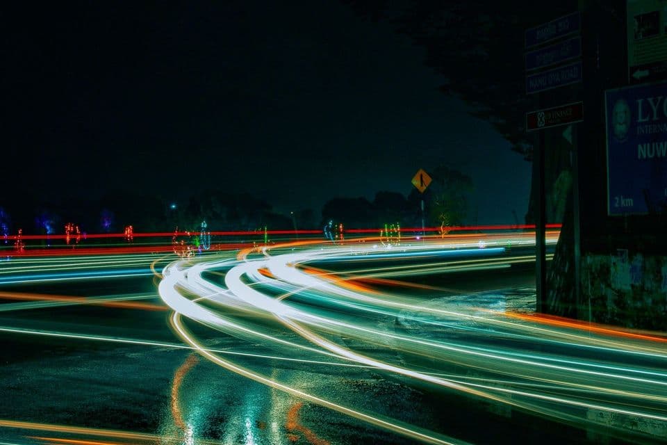 Long-exposure photograph of a wet road at night, white, orange and teal light trails curving through the frame past a road sign reading NANU OYA ROAD, no vehicles visible