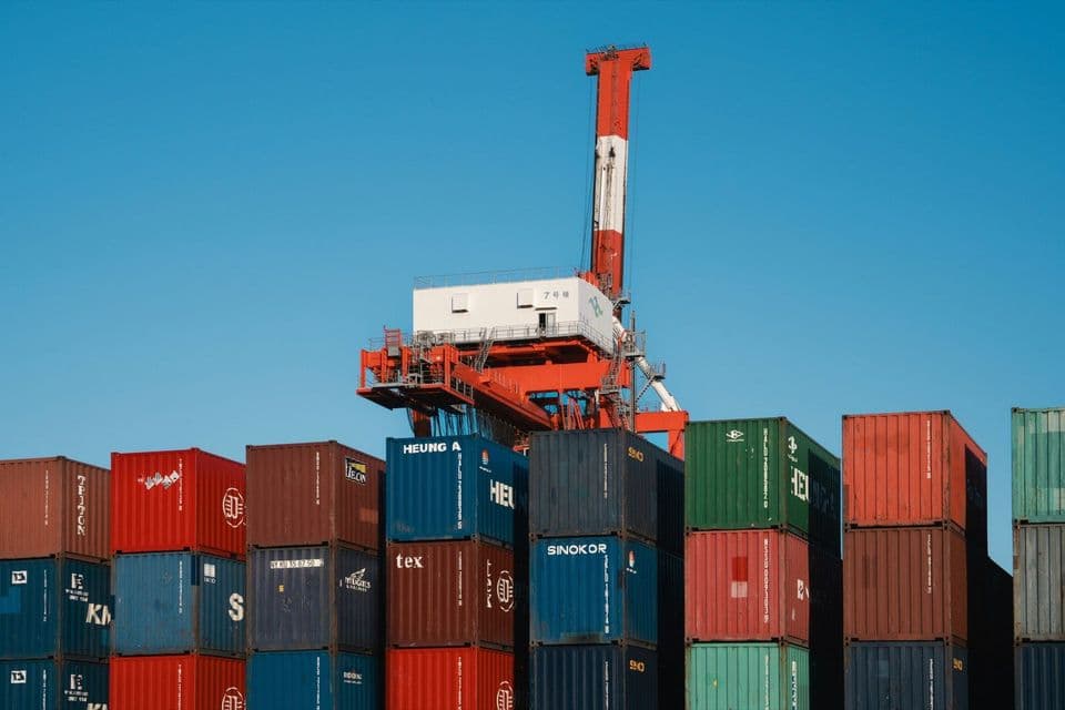 Shipping containers stacked several high in red, blue and green rows at a port, an orange gantry crane rising behind them against a clear blue sky