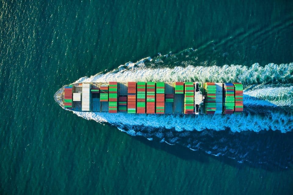 A container ship photographed from directly above, its deck stacked with differently coloured freight containers, cutting a wake across open water