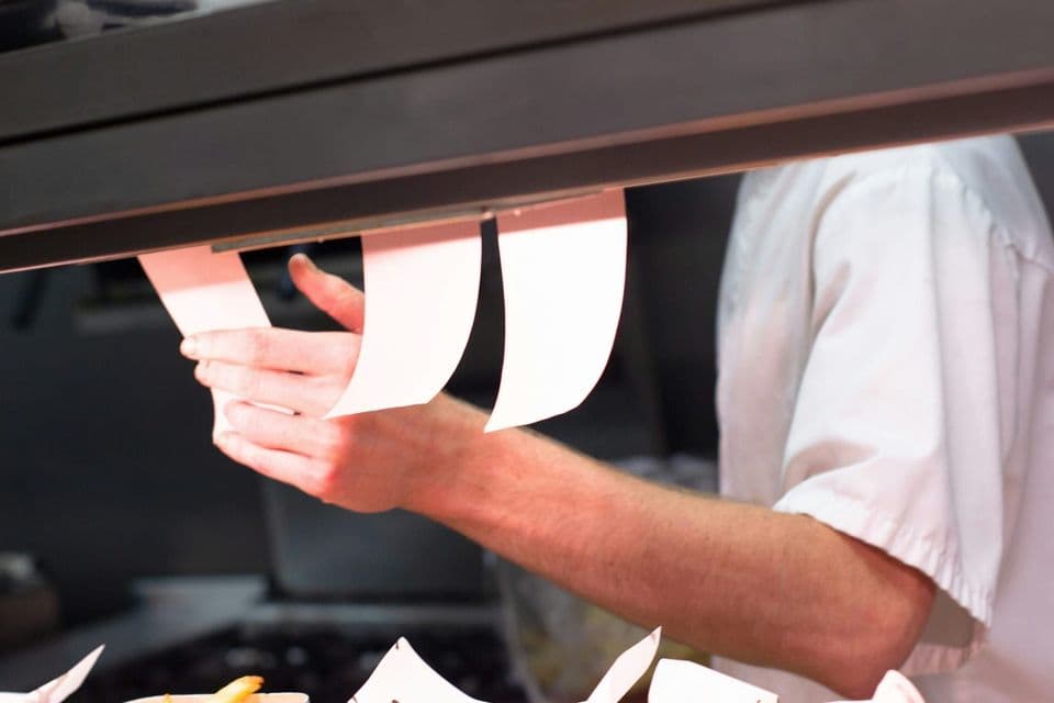 A cook reaching for one of several paper order tickets clipped to a rail under a heat lamp in a restaurant kitchen