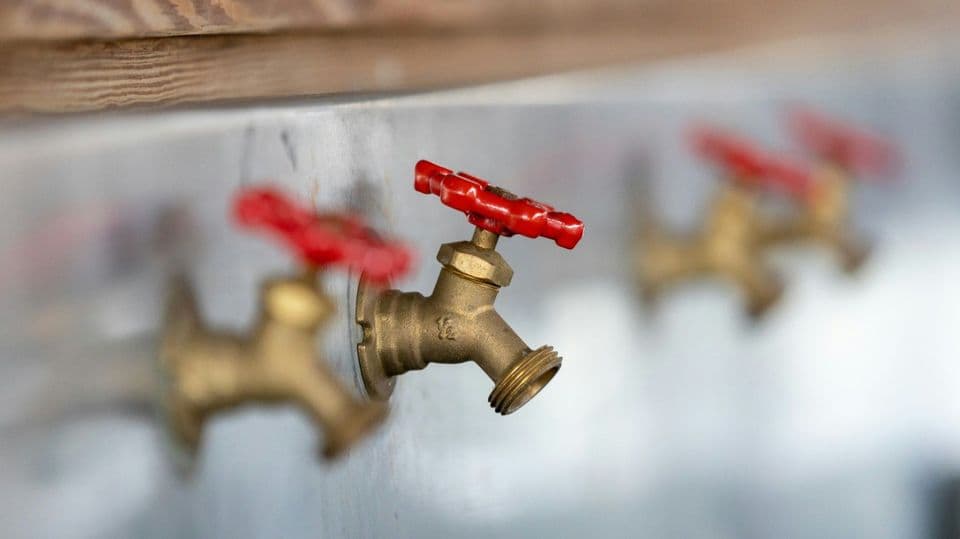 A row of brass wall taps with red handwheels mounted along a metal wall, the second one sharp and the others out of focus