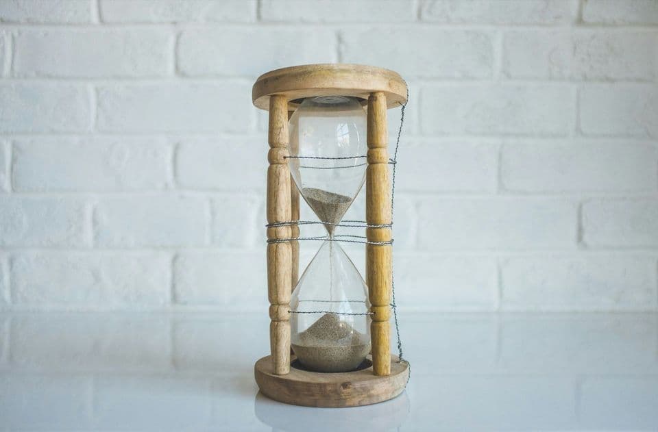 A wooden-framed hourglass standing on a white surface against a white brick wall, sand part-way through falling into the lower bulb