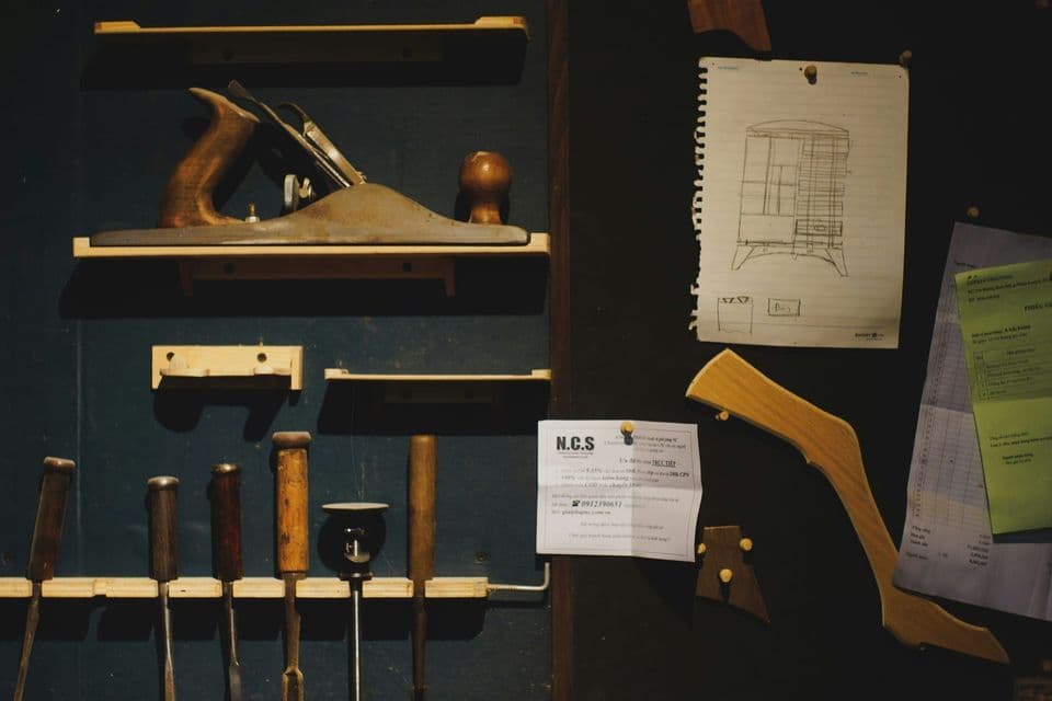 A workshop wall with a hand plane on a shelf and chisels in a rack, next to a hand-drawn furniture plan and order sheets pinned up beside them