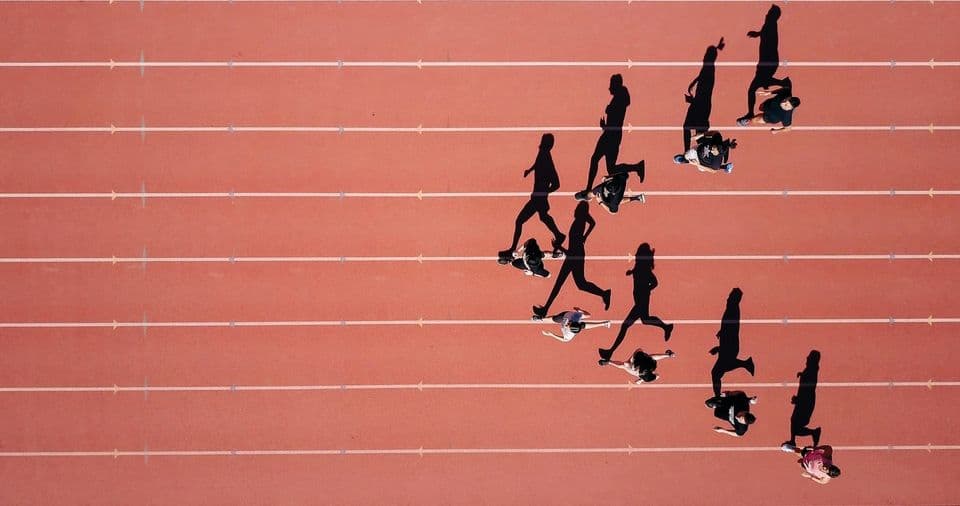 Runners photographed from directly above on a red running track, strung out in a diagonal line across the lanes with long shadows behind them