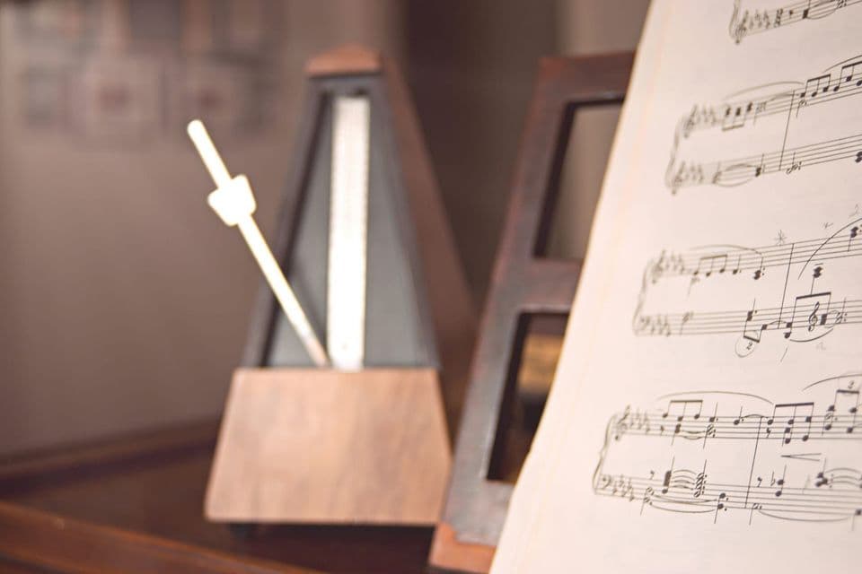 A wooden metronome standing on a piano with its pendulum arm mid-swing, an open page of handwritten sheet music sharp in the foreground
