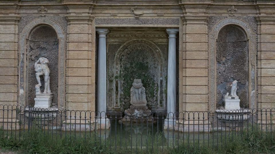 A weathered stone fountain wall with marble statues standing in its niches, the figure on the right reduced to a headless and armless torso on its plinth, an iron fence across the front