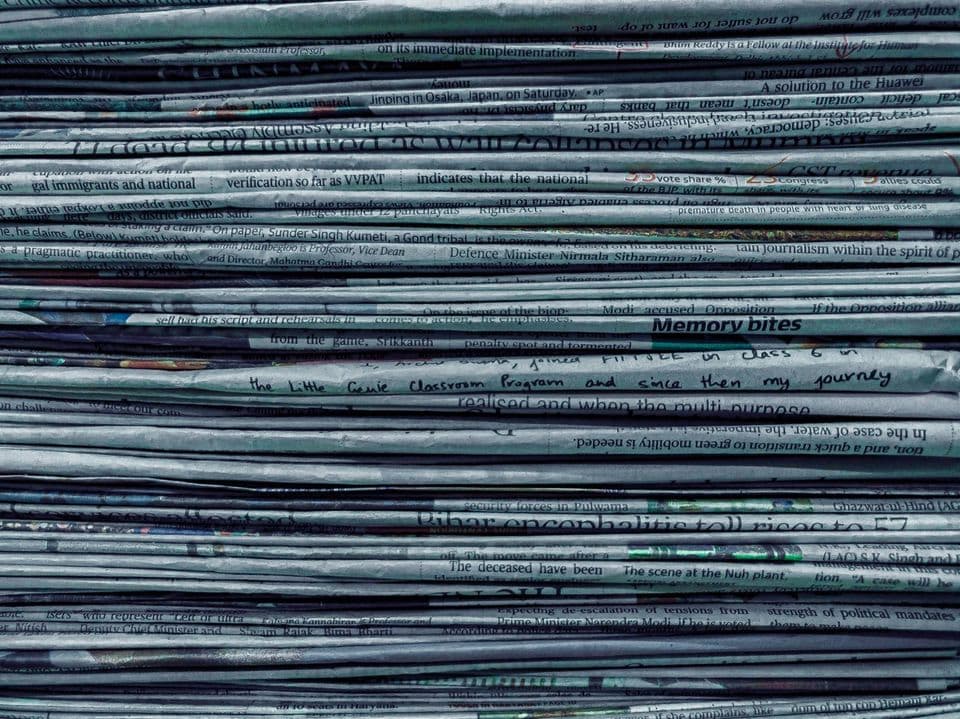 A tall stack of folded newspapers photographed edge-on, headlines running along every layer, tinted blue-green