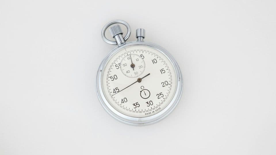 A chrome mechanical stopwatch with a white dial lying on a plain light background, hand stopped between 45 and 50 seconds