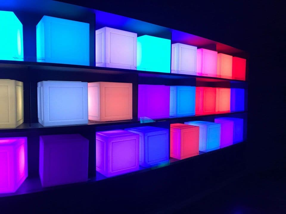 Three shelves of identical illuminated cubes in a dark room, each cube glowing a different colour, blue, white, pink and violet, repeating across the grid