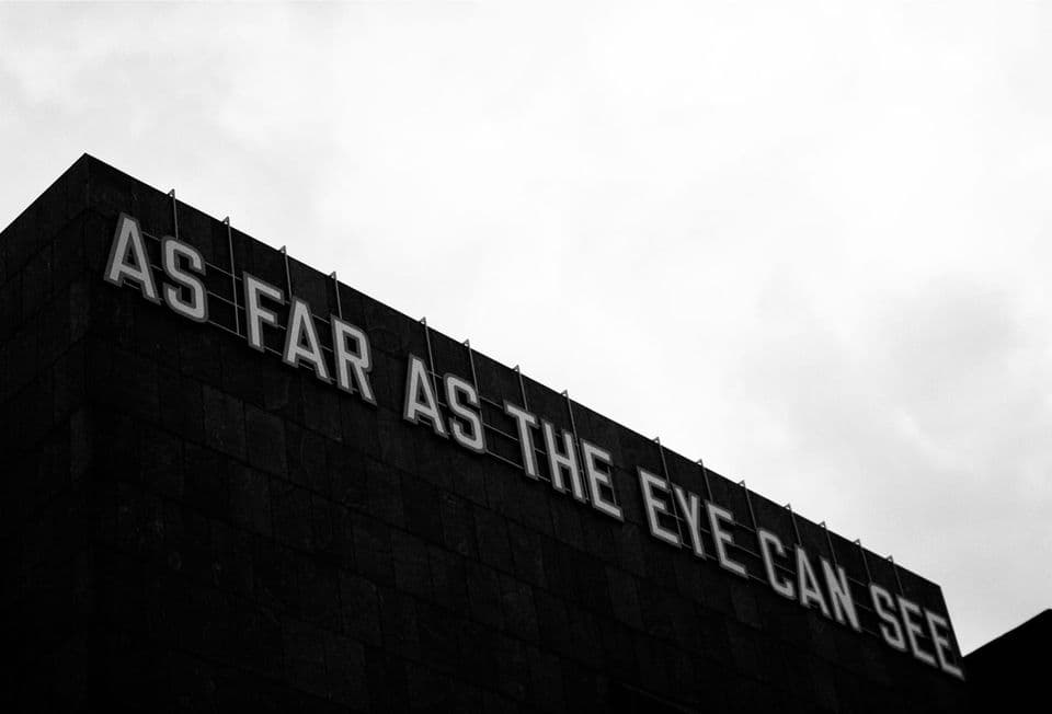 The words AS FAR AS THE EYE CAN SEE mounted in large letters along the roofline of a dark building against a blank white sky