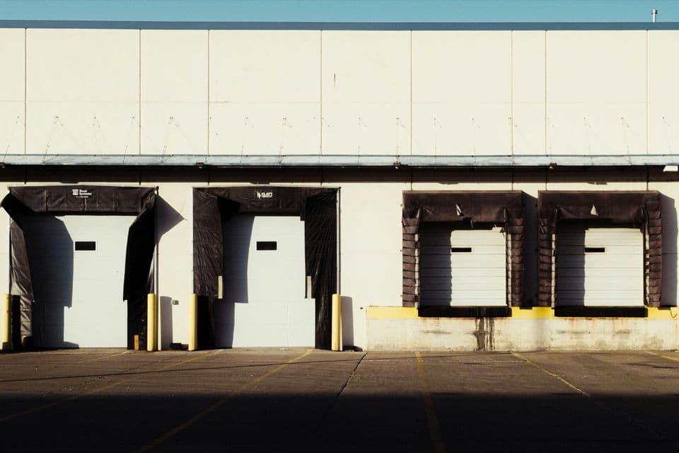 Four loading bays along the side of a pale warehouse building, roller shutter doors closed behind black rubber dock seals, the concrete apron in front of them empty