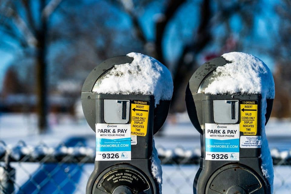 Two identical coin-operated parking meters side by side outdoors, both capped with snow, Park and Pay stickers on their fronts and a snow-covered park blurred behind them