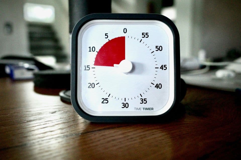 A black Time Timer countdown clock standing on a wooden desk, its red disc filling the segment between 0 and 15 minutes on the white dial, the room behind it out of focus