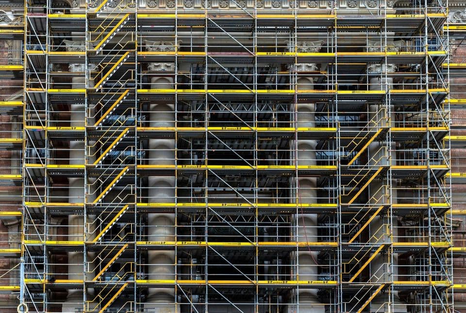 Metal scaffolding with yellow platforms and stairs covering the columned stone facade of a building