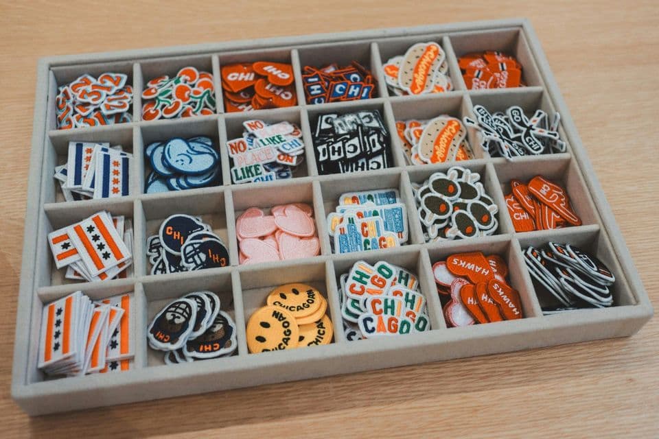 A felt-lined sorting tray on a wooden desk, each compartment holding a different embroidered patch design