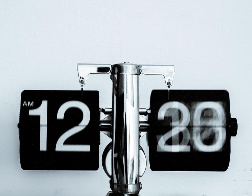 A retro flip clock in black and white against a white wall, showing 12:20 AM with the minute card caught mid-flip