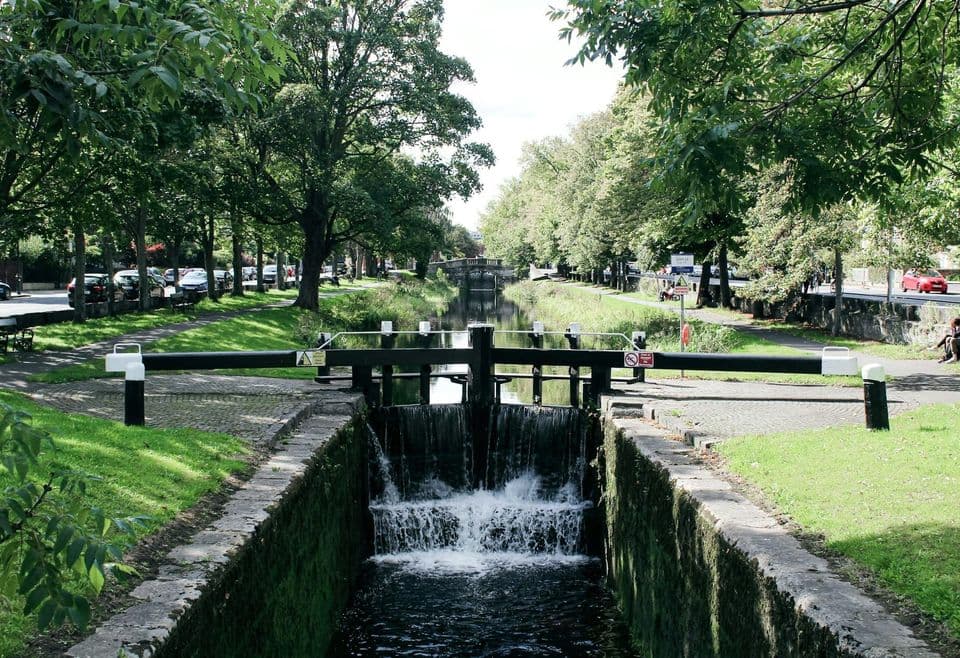 Closed black and white canal lock gates with water spilling through into the lower chamber, a tree-lined canal running into the distance behind them