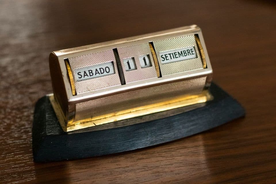 A small brass mechanical desk calendar on a dark wooden table, its rotating bands set to SABADO 11 SETIEMBRE