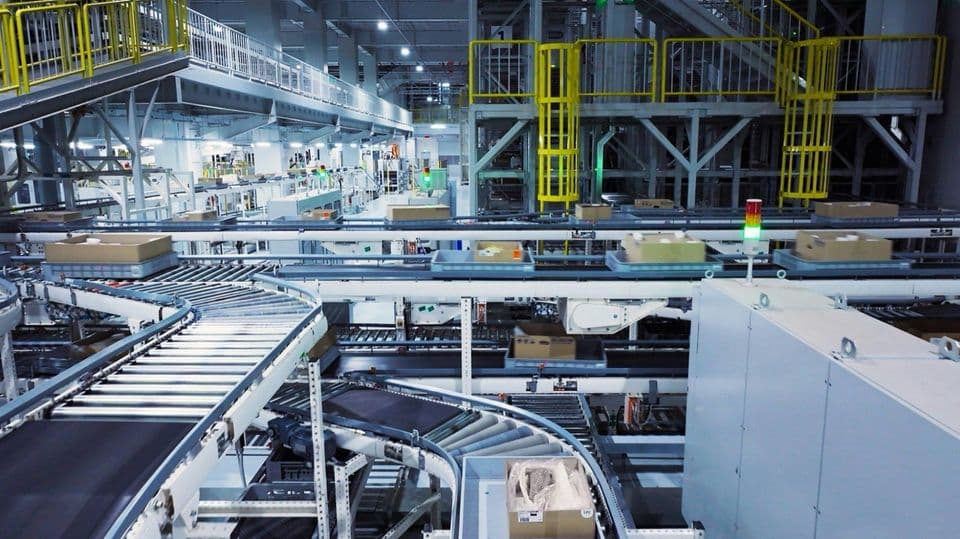 An automated warehouse sorting hall seen from a gantry, cardboard boxes and plastic totes riding several levels of roller conveyors, a green and amber stack light standing on the near line