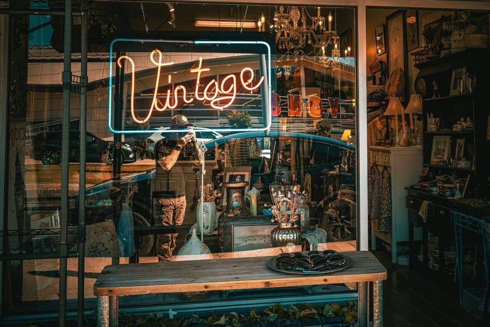 The window of a vintage shop at dusk, a neon Vintage sign glowing behind the glass above a table of second-hand goods